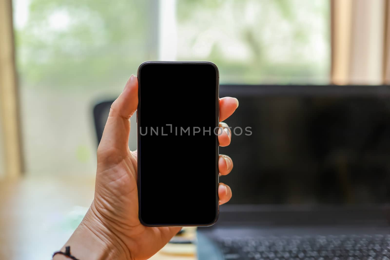 Close up of man hand holding mobile smartphone with blank screen copy space and computer laptop in office.