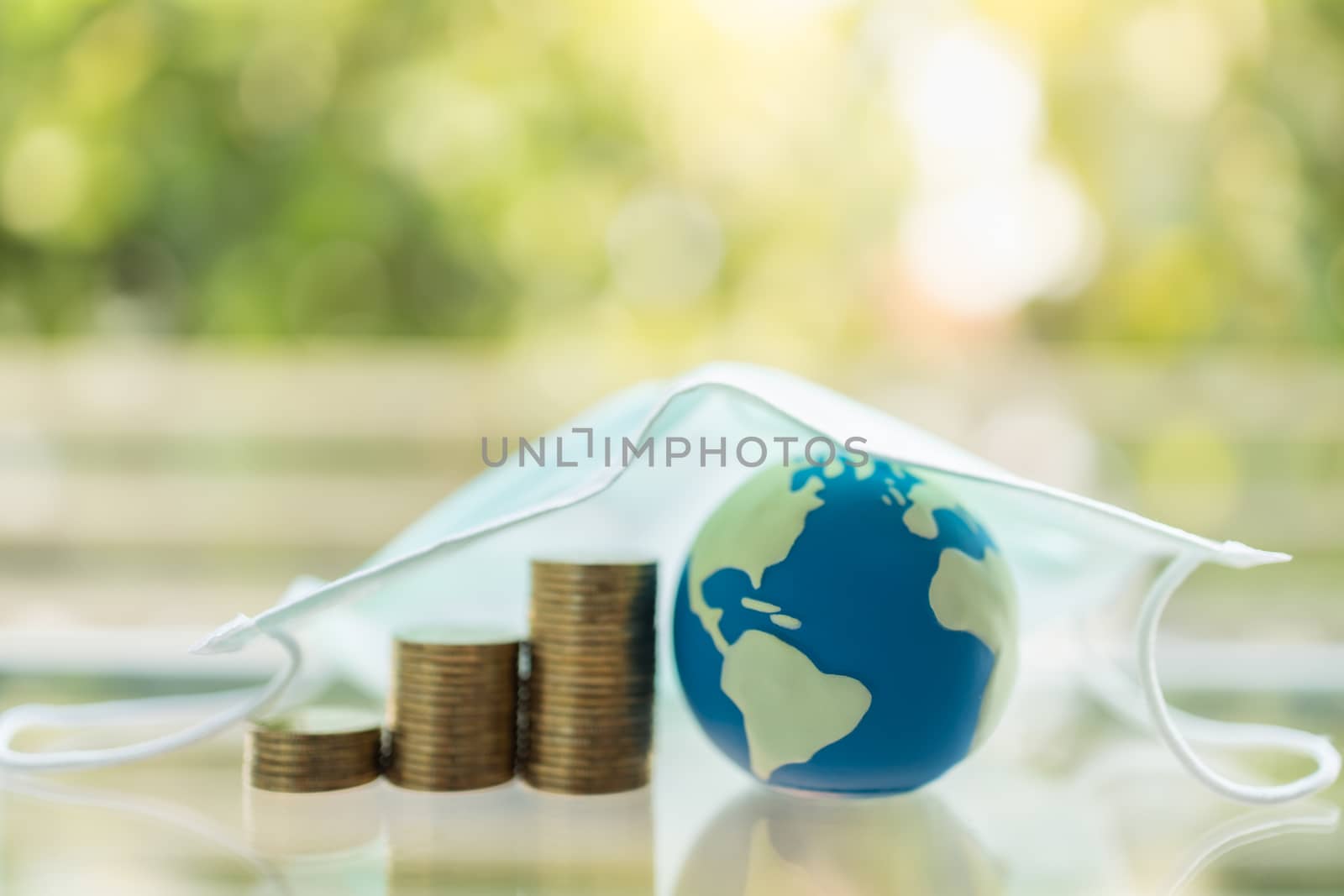 Global Money, Business, Healthcare in Cornavirus (COVID-19) Situation Concept. Mini world ball with stack of gold coins under surgical face mask and green nature background with copy space.