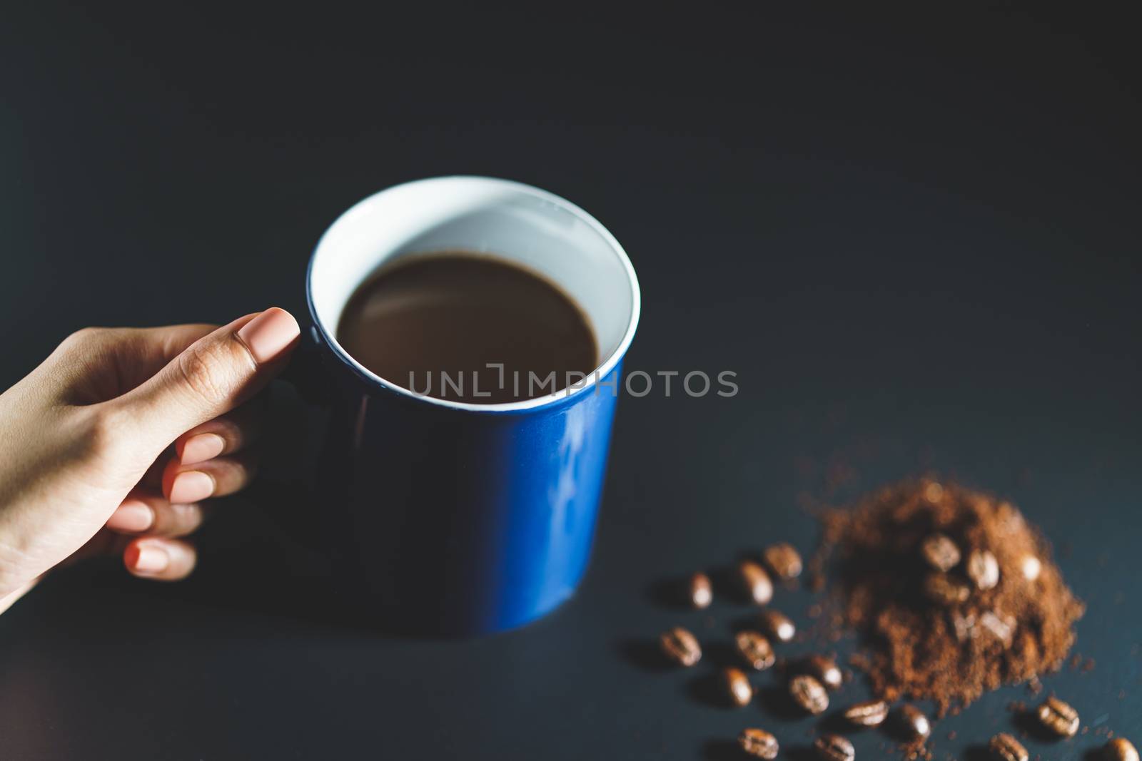 Hand holding a cup of coffee with coffee beans and powder on black background.