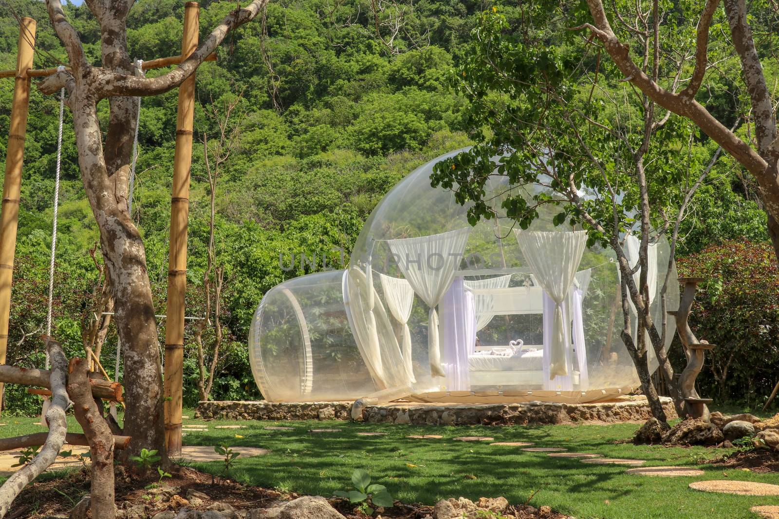 Romantic bubble house with transparent walls. White wooden double bed with mosquito net inside bubble. Honeymoon in an inflatable tent. Tourist attraction in tropical paradise on Bali island in Indonesia.