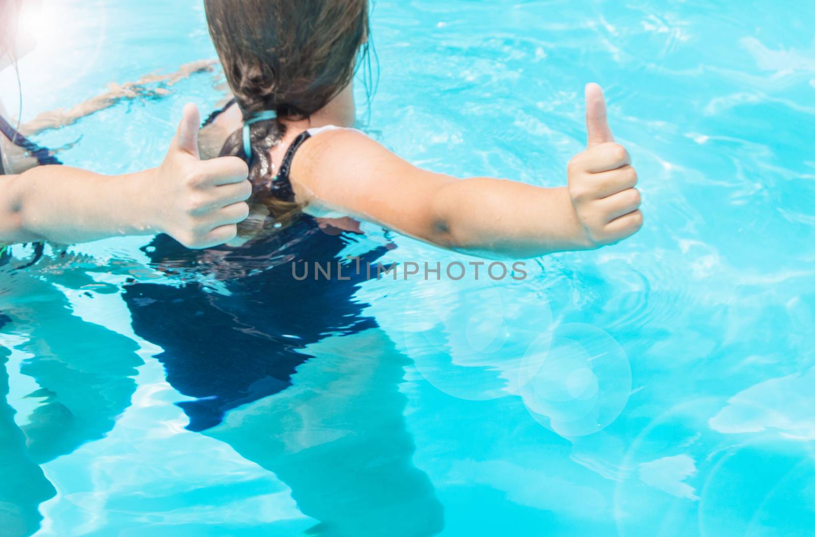 Thumbs up two teenage girls with long hair in swimsuits in a pool with blue sparkling water in the open, sunlight, glare summer.