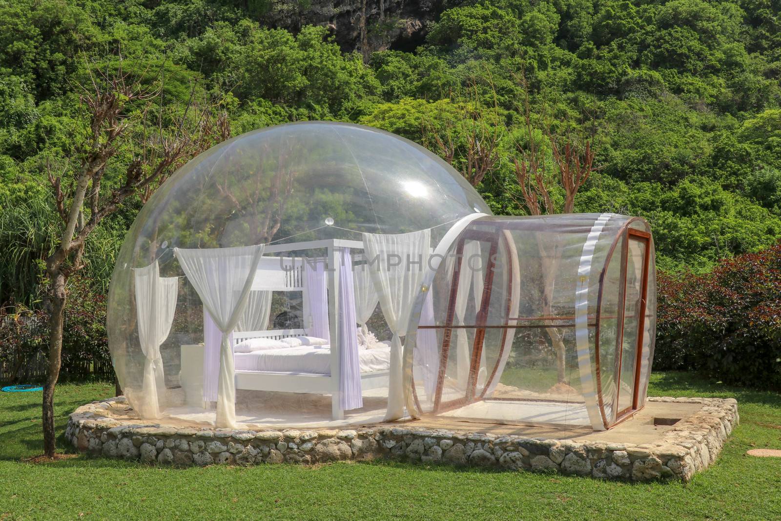 Glamping on Bali beach, in bubble house with transparent walls. White wooden double bed with mosquito net inside bubble. Honeymoon in an inflatable tent. Tourist attraction in tropical paradise.