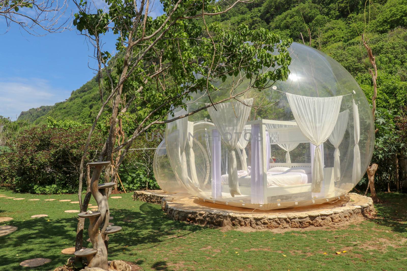 Bubble house with transparent walls. White wooden double bed with mosquito net inside bubble. Honeymoon in inflatable tent for tourists. Bubble hotel in tropical paradise on Bali island in Indonesia.