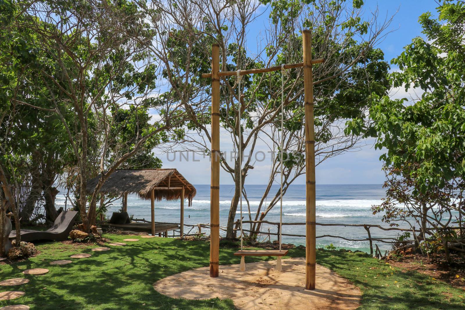 A swing made of bamboo trunks and white ropes on the coast of Bali island. Romantic beachfront location with fine sand. Tropical garden with English lawn. Azure blue sky and Indian ocean on background