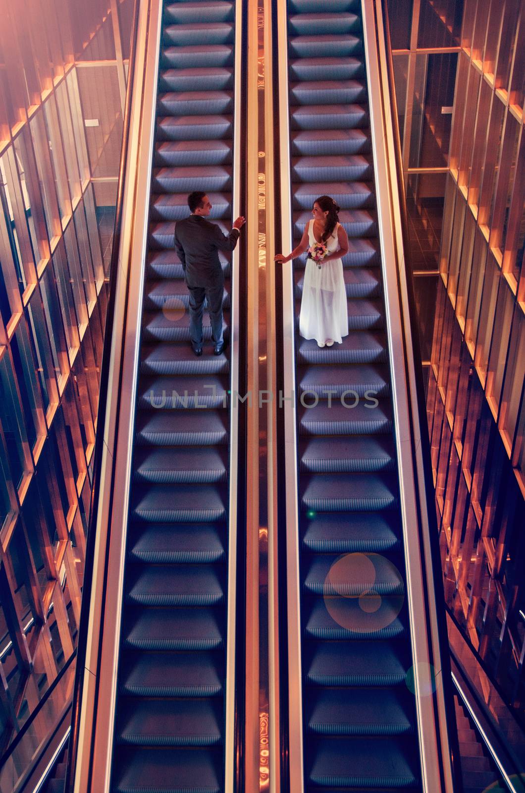 Smiling bride and groom on the rise escalators are located in parallel, drawn to each other hands. by Peruphotoart