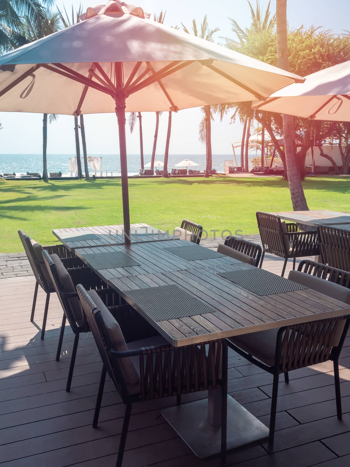 Empty wooden dining table and chairs with beach umbrella near the green yard with sea view. Outdoor  breakfast area in hotel, vertical style.