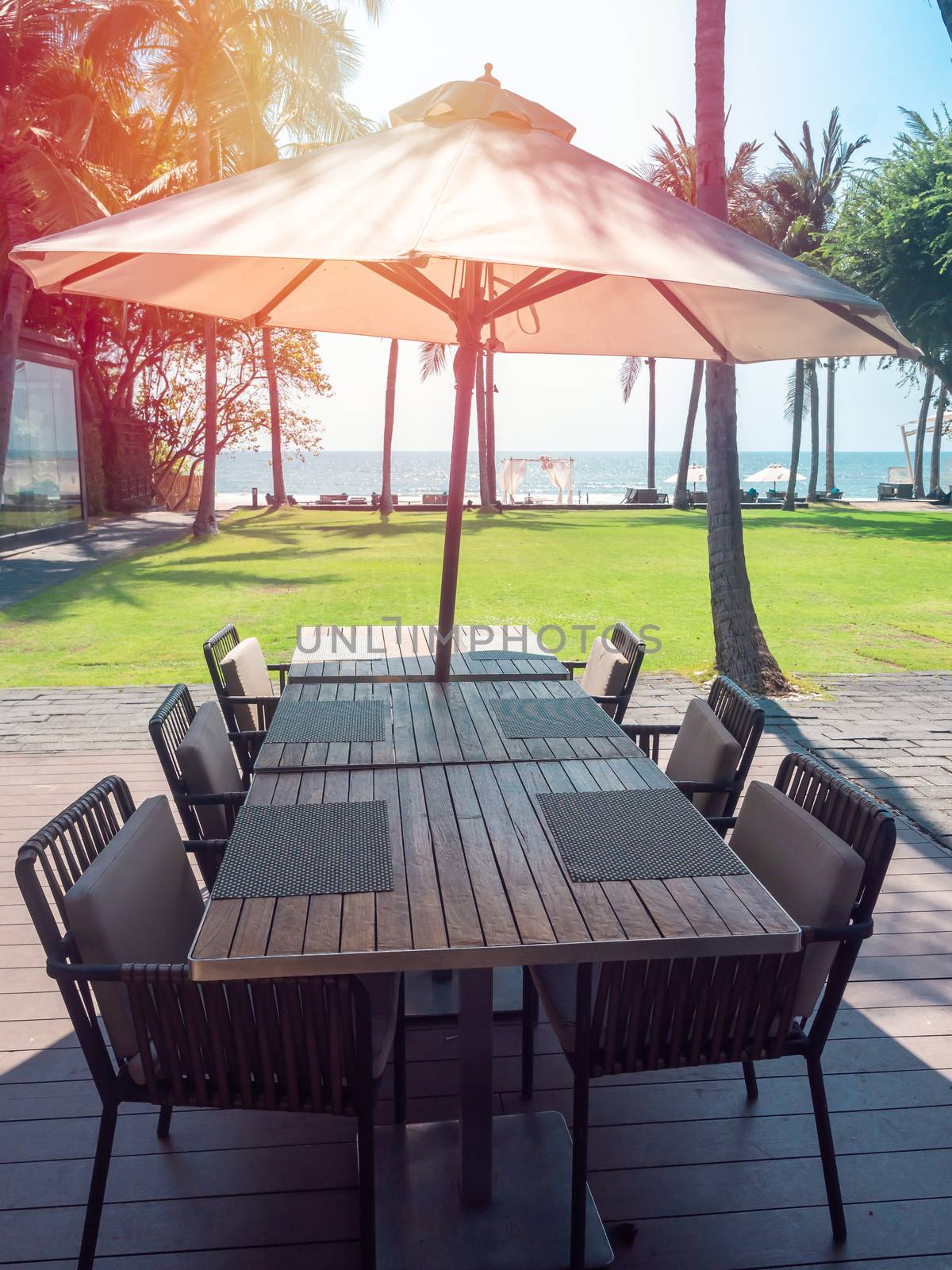 Empty wooden dining table and chairs with beach umbrella near the green yard with sea view. Outdoor  breakfast area in hotel, vertical style.