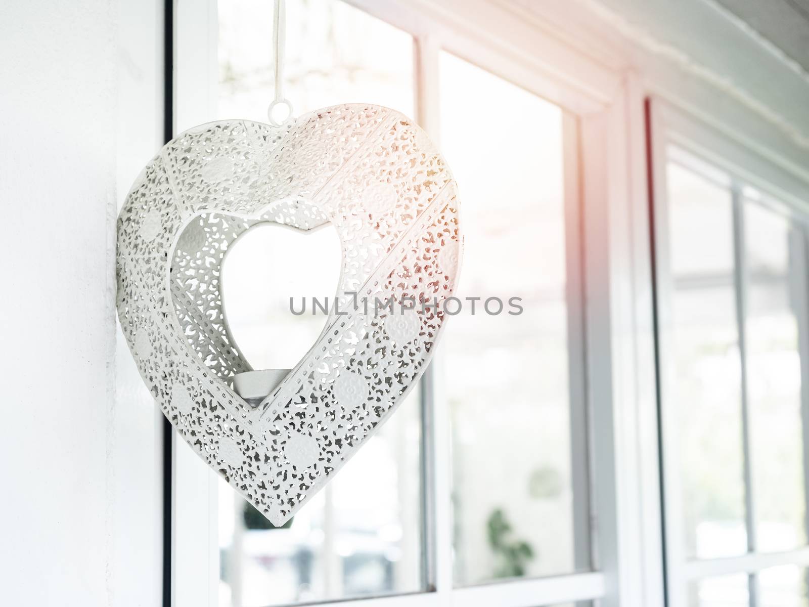White beautiful vintage iron heart shaped lamp hanging on the white wall near the glass window.