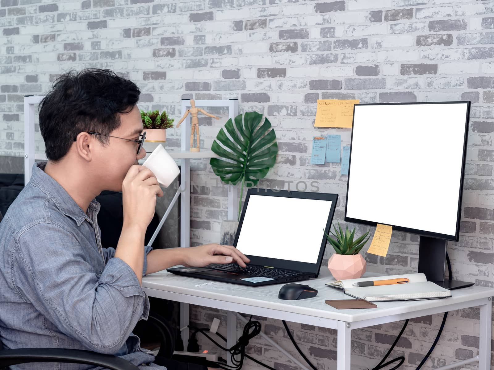 Asian man drinking coffee and working with laptop computer with white blank screen and watching another monitor in his room, condominium. Work at home and business online concept.