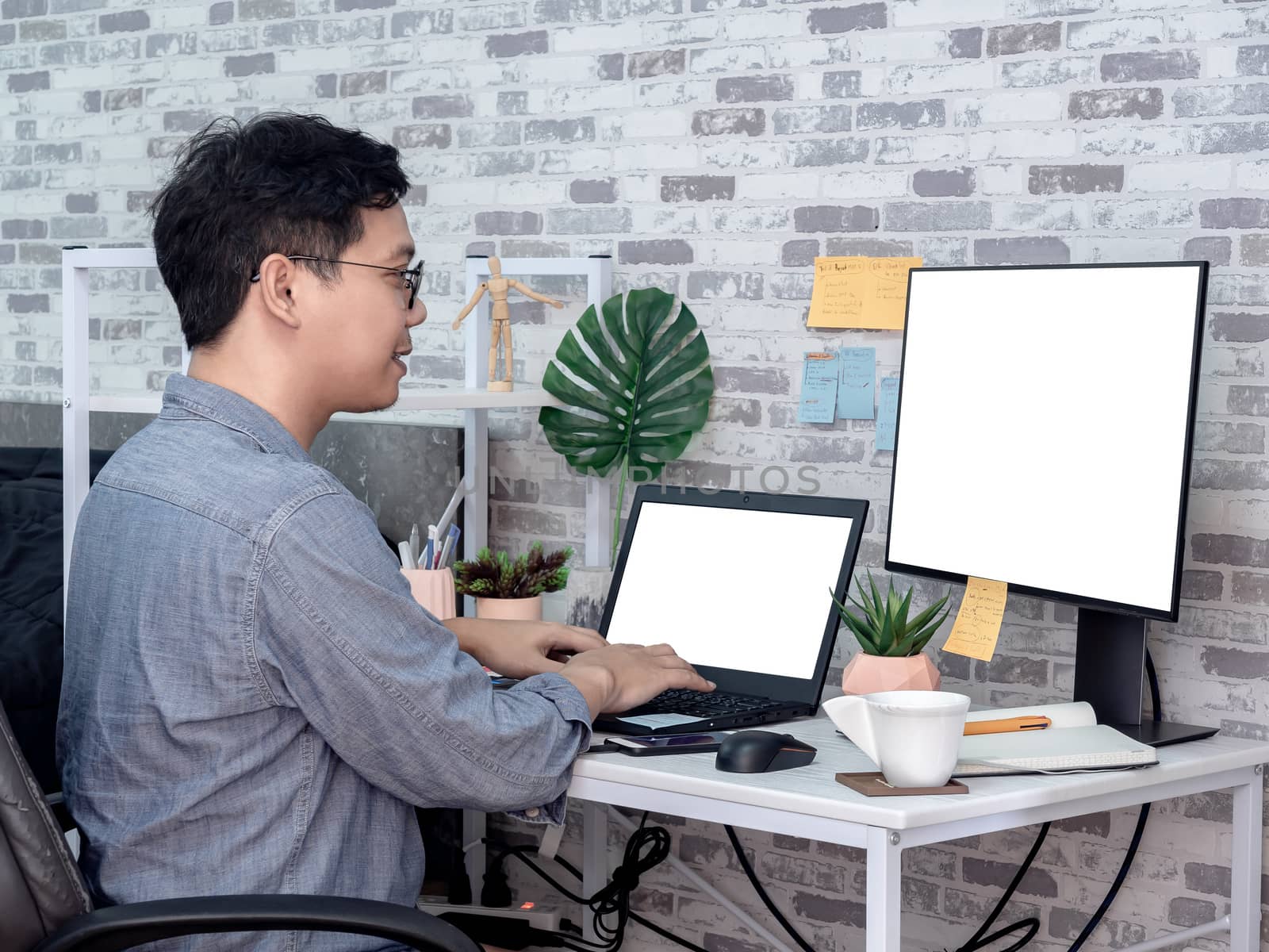 Asian man working and typing with laptop computer with white blank screen and watching another monitor in his room, condominium. Work at home and business online concept.