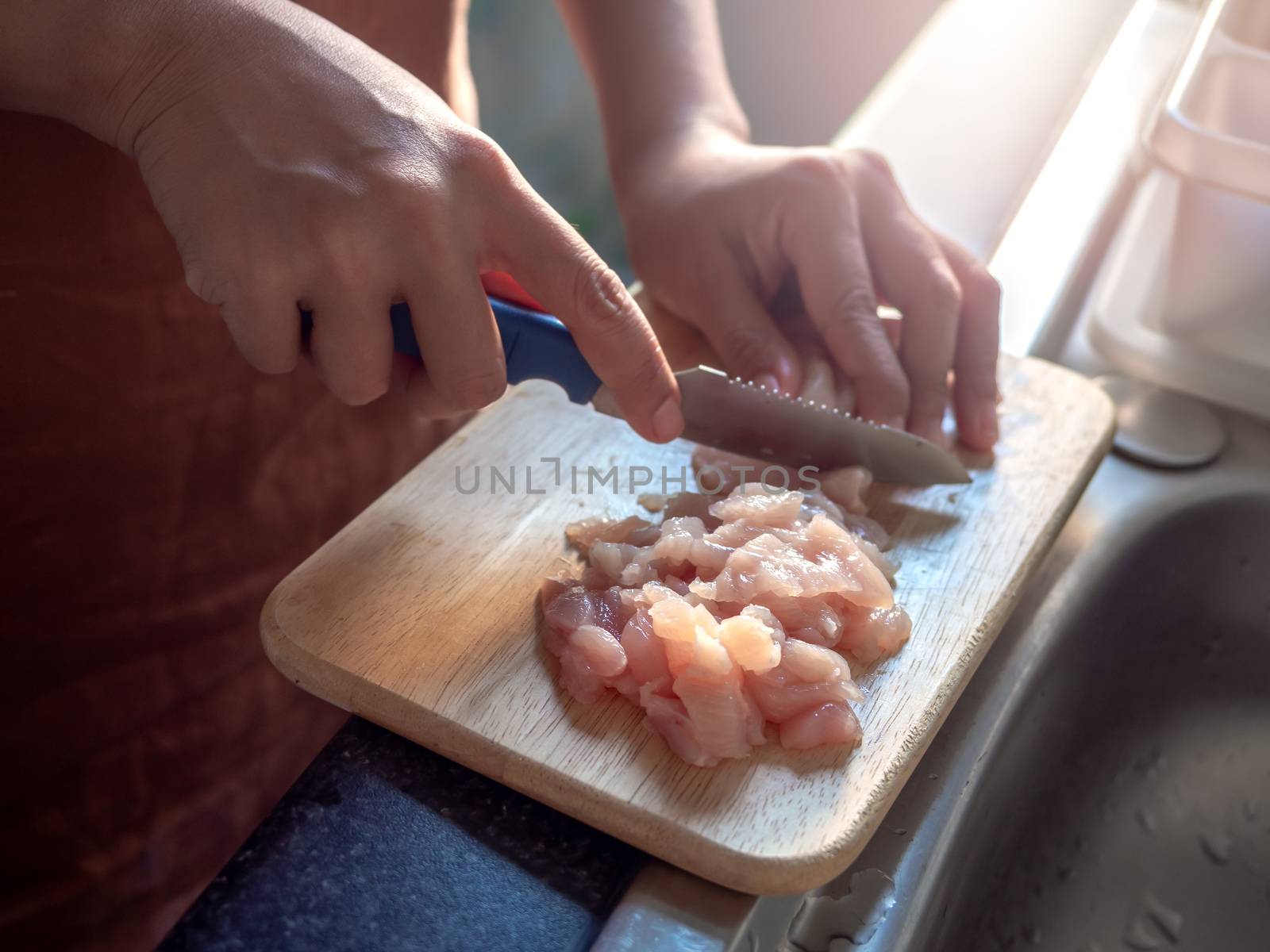 Close-up hand using a sharp knife, slice the chicken on a wooden cutting board near the window in room in condominium in the evening. Cooking at home.