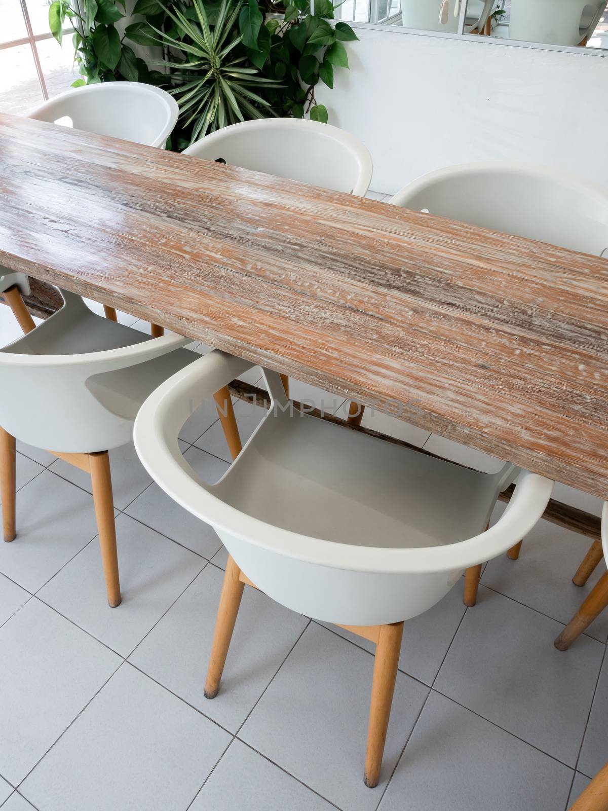 Table set, white modern plastic armchairs and empty wooden table near green leaves in cafe, vertical style.