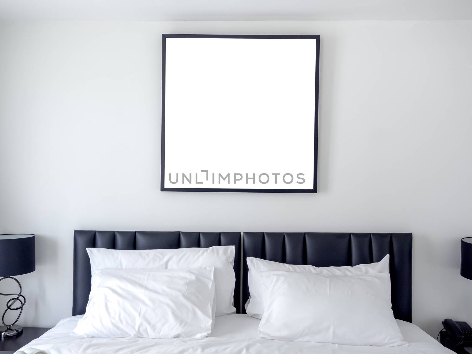 Blank photo square frame mockup on white wall above the bed with white clean pillows in the bedroom.