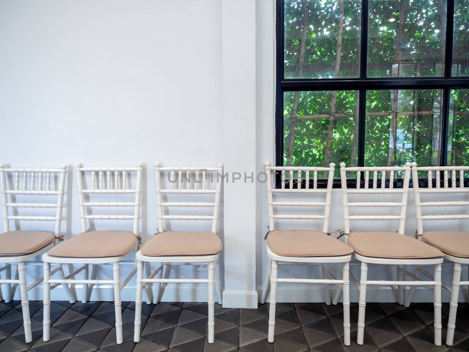 Row of clean white vintage wooden chairs with pads on modern stone tiles floor on white wall and glass window with green garden background.