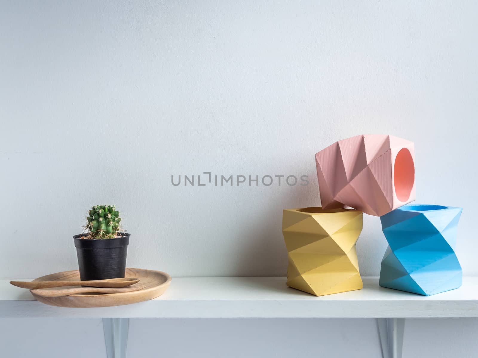 Cactus pot. Concrete pot. Three empty blue, pink and yellow modern geometric concrete planters and green cactus on wooden dish on white wooden shelf isolated on white background.