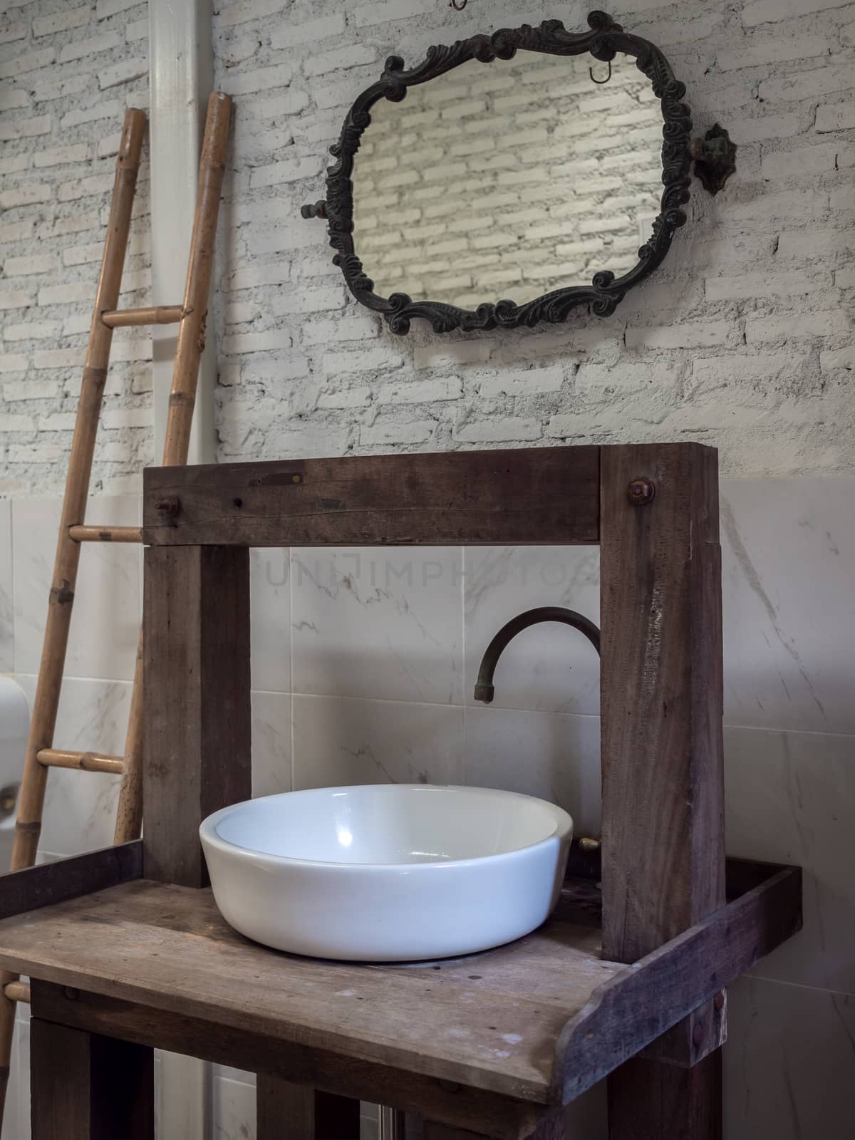 White clean ceramic sink bath and faucet on vintage wooden table and vintage mirror and wood ladder decoration on white brick wall background vertical style.