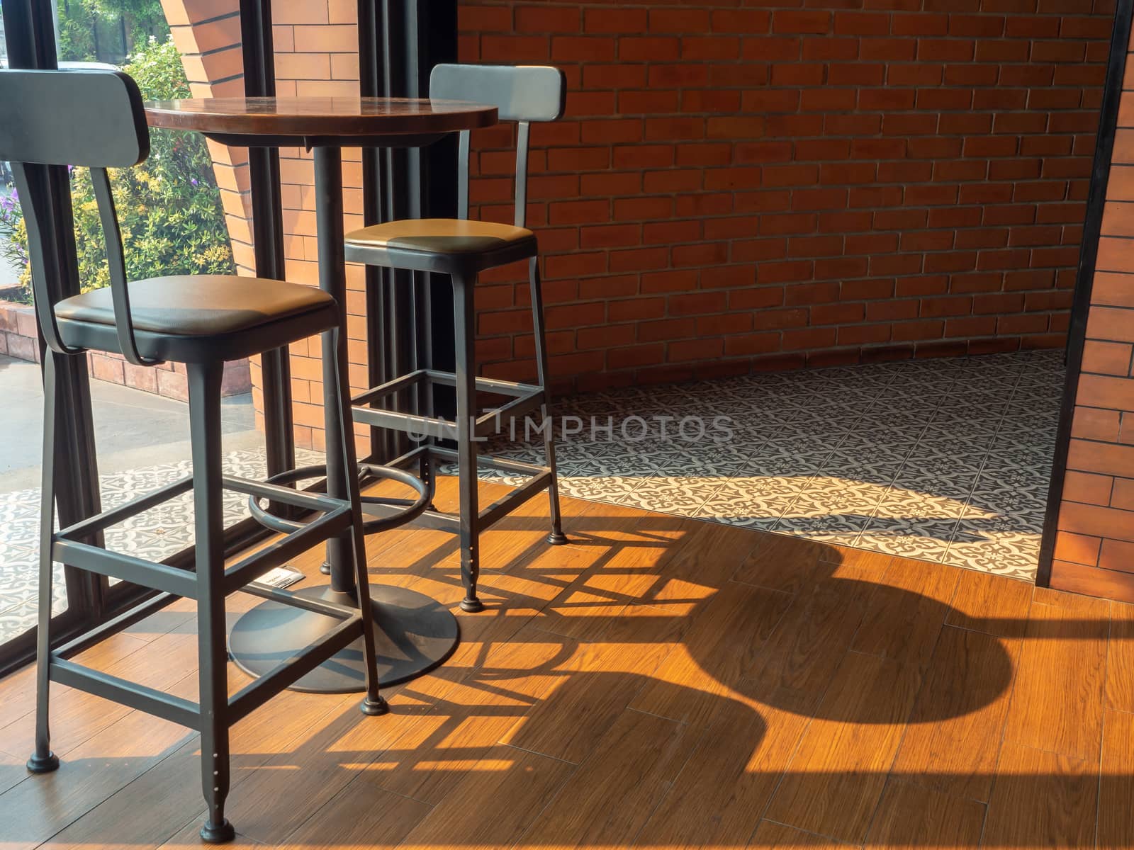 Cafe decoration design. Round wooden table with two modern bar stools on wooden floor near the glass window near brick wall on sunshine day.