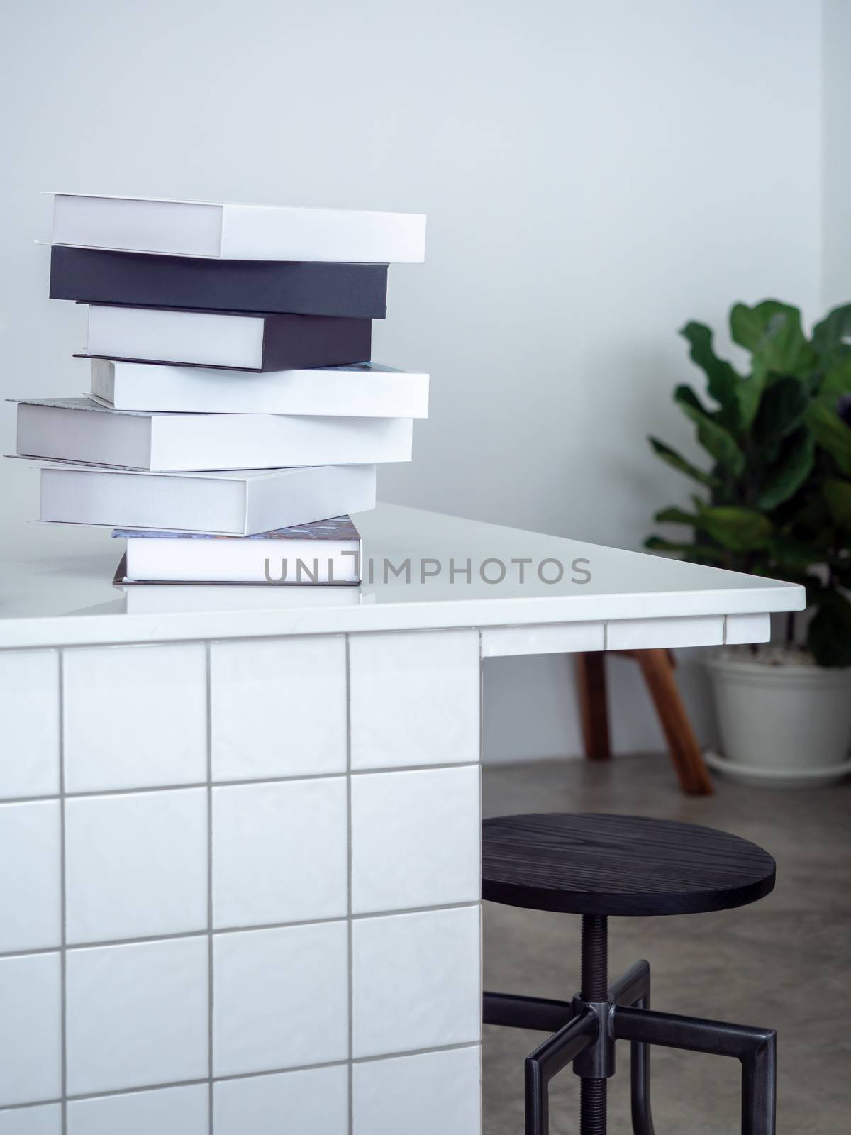 stack of black and white mockup books on white counter bar with wooden bar stool and green leaves in pot in white cafe background minimal style, vertical style.