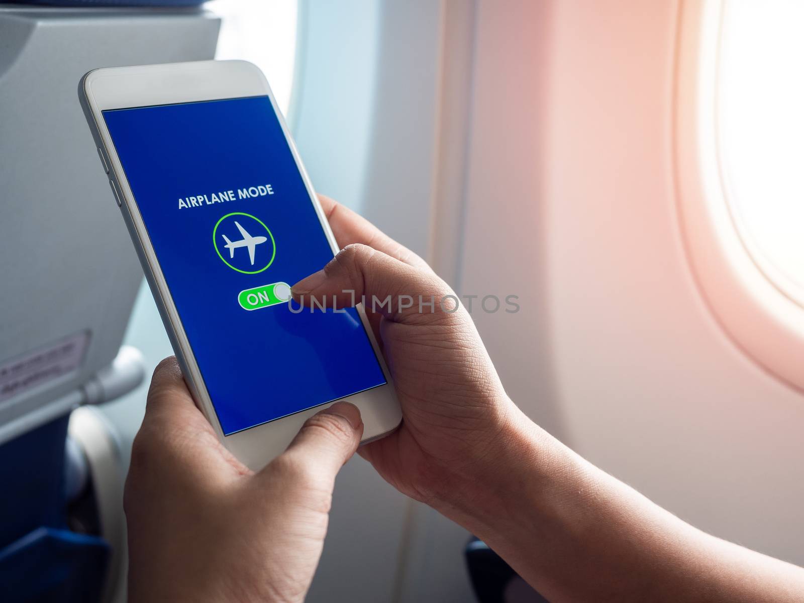 Flight mode concept. Hand holding white smartphone and turned on airplane mode on screen near the window on the airplane with copy space.