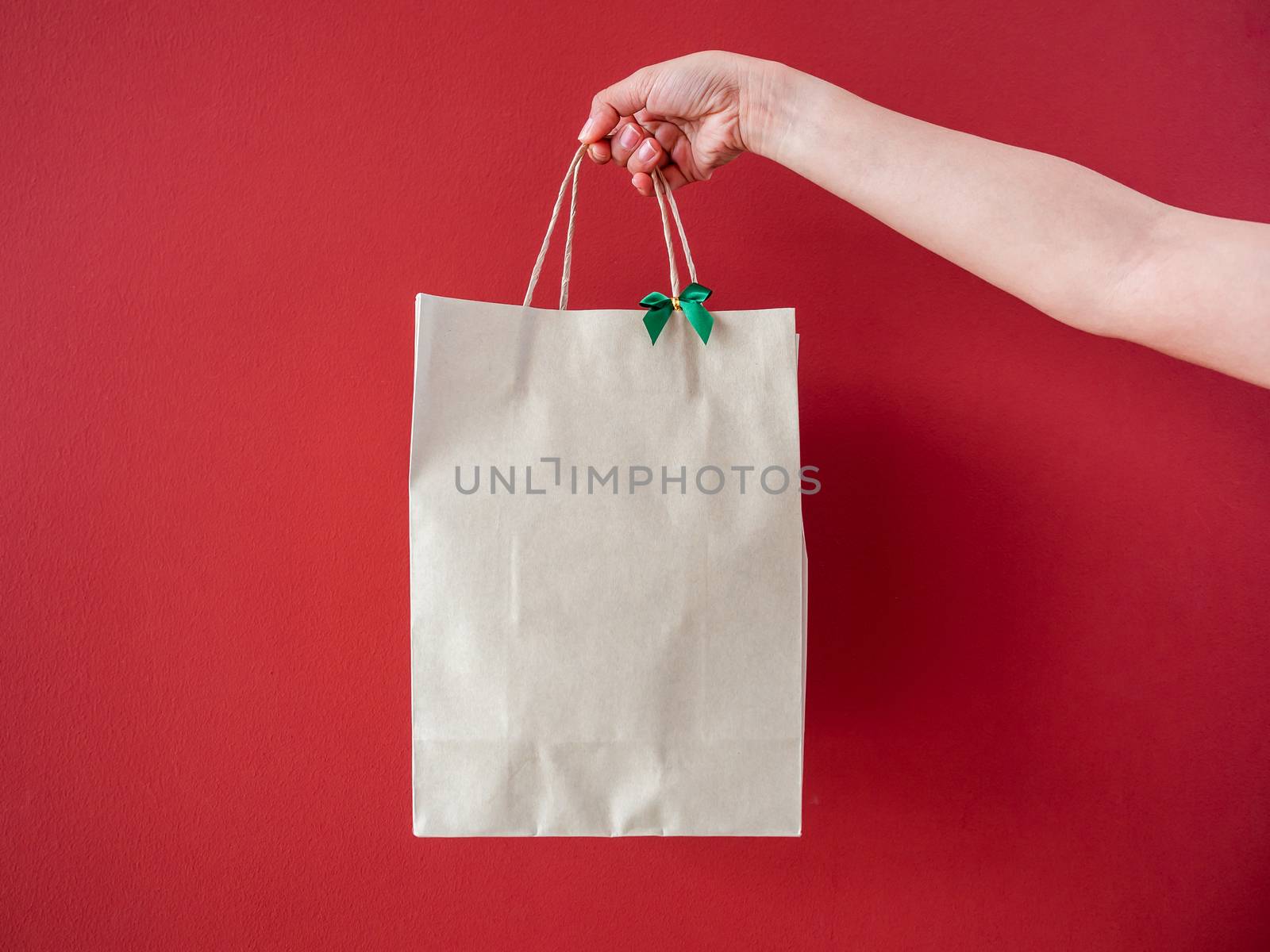 Woman's hand holding blank recycled paper bag decorate with little green bow on red wall background.