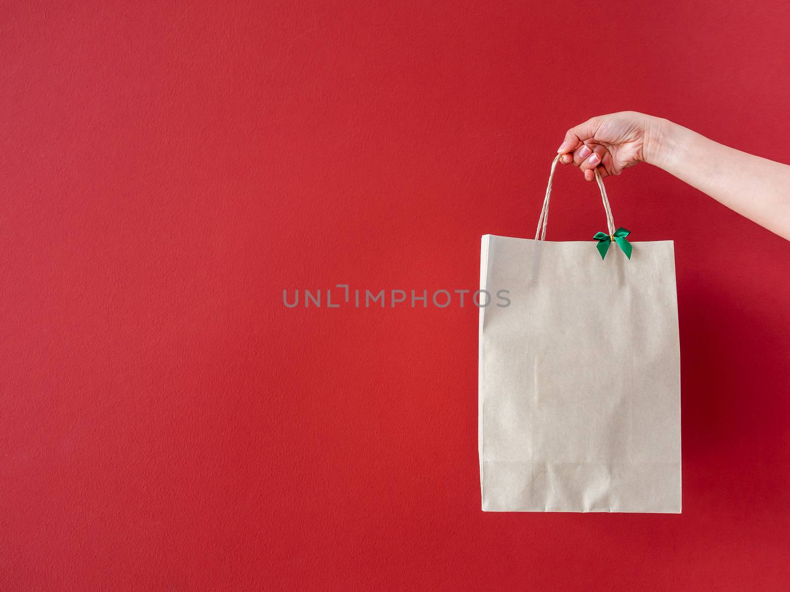 Woman's hand holding blank recycled paper bag decorate with little green bow on red wall background with copy space.