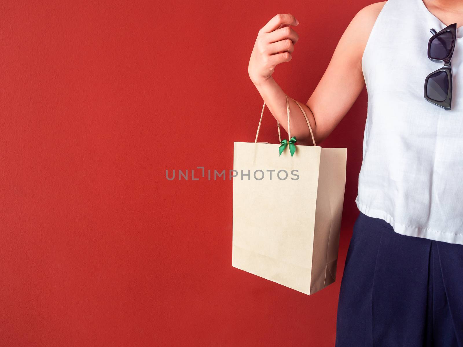 Woman's hand holding blank recycled paper bag decorate with little green bow on red wall background with copy space.
