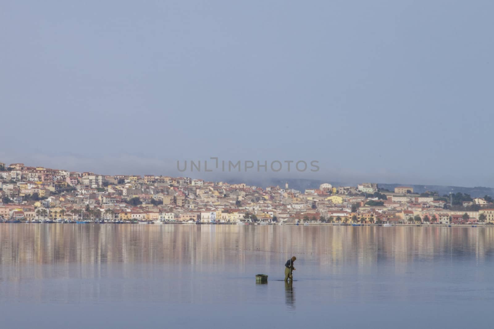 A fisherman at work bending in the flat sea water that reflects the houses of Sant'Antioco in Sardinia