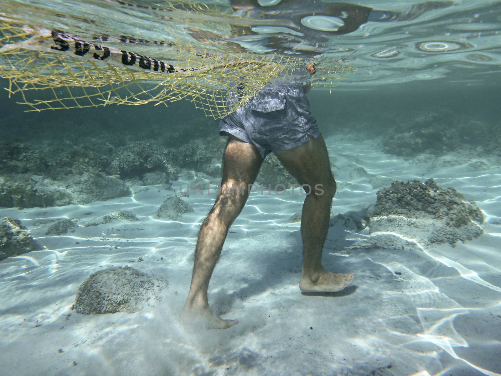 Plastic pollution in the ocean and in the sea: underwater shot of a man walking on the seabed dragging a fishing net and a piece of plastic in the crystal clear water