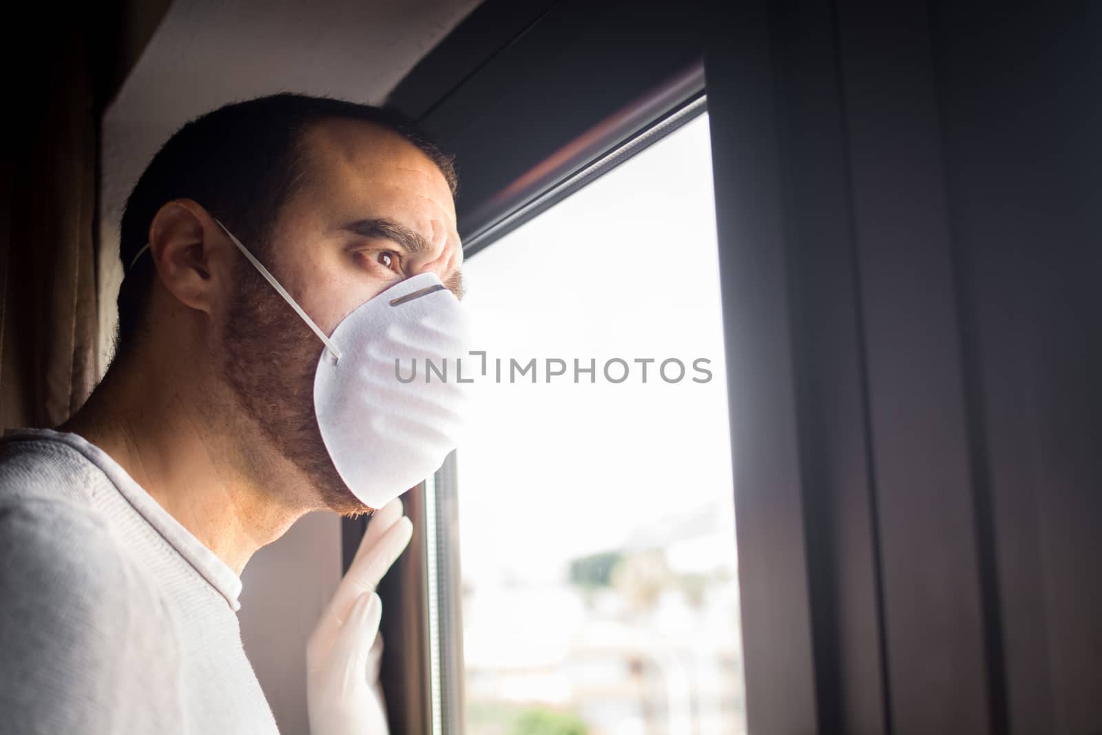 Man with face mask and gloves looking out the window. Stay at home concept.