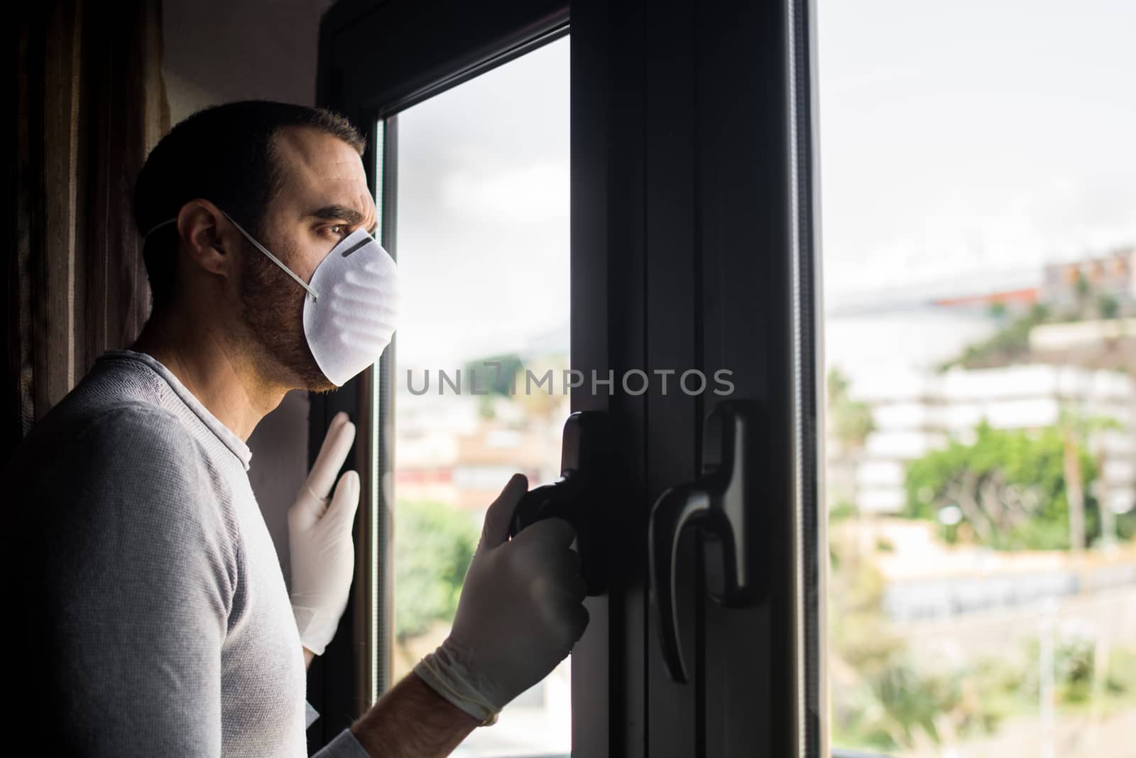 Man with face mask and gloves looking out the window. Stay at home concept.