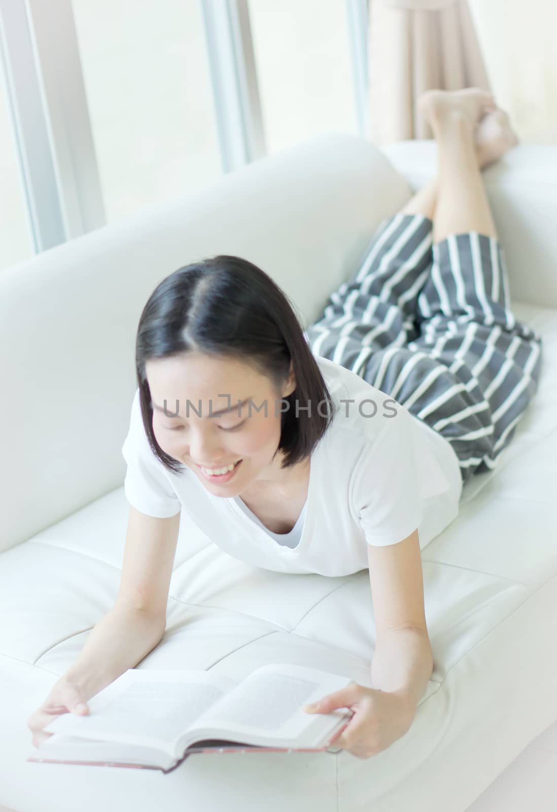 Happy young woman reading storybook on couch at home by ekachailo