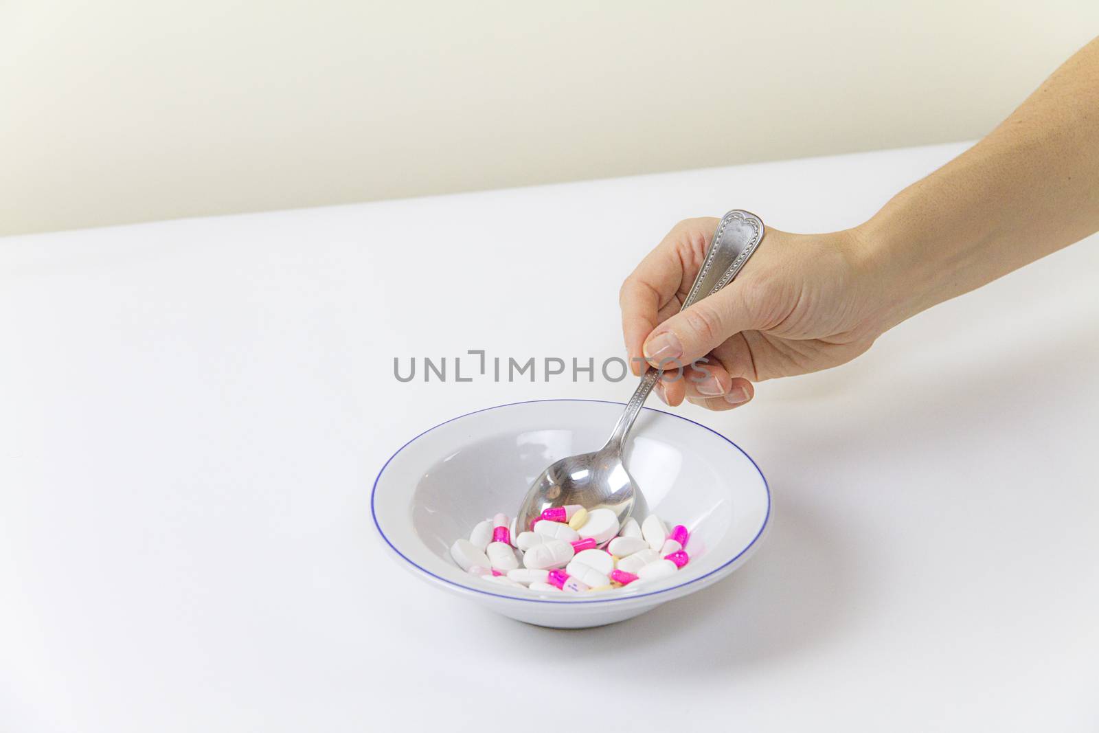 Addiction to medicines and drugs in modern times: a hand of a young woman with a spoonful of tablets taken from a plate full of medicines on white table and background