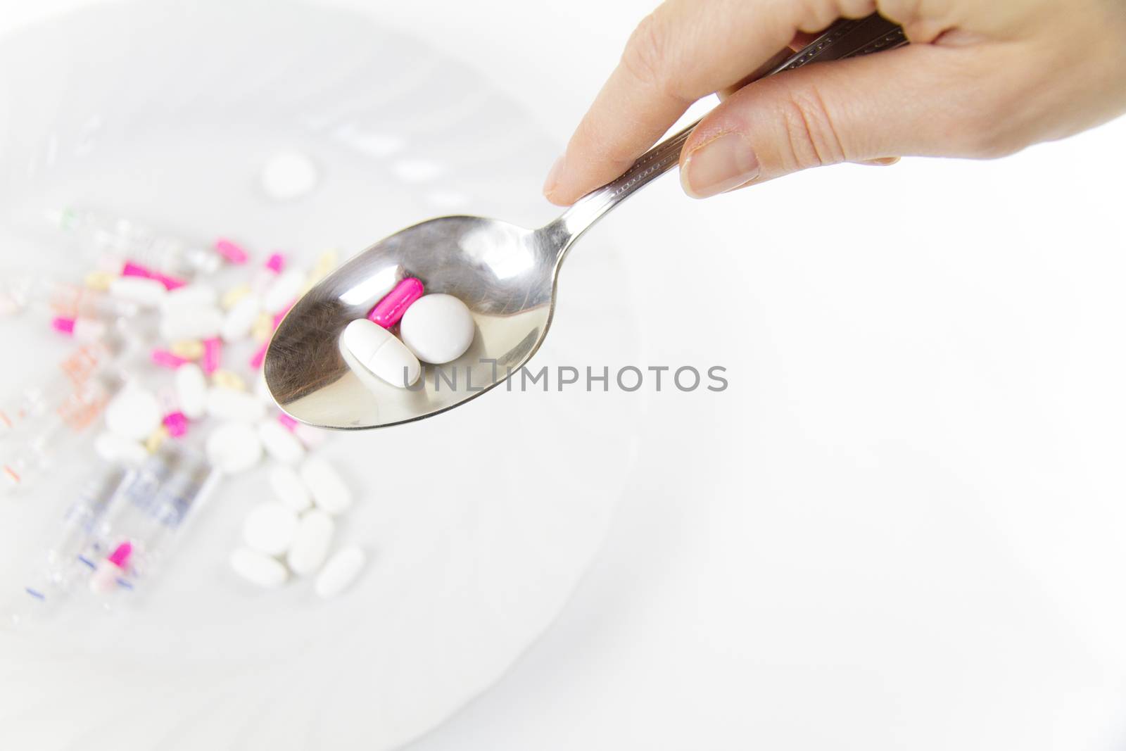 Addiction to medicines and drugs in modern times: a hand of a young woman with a spoonful of tablets taken from a plate full of medicines on white table and background