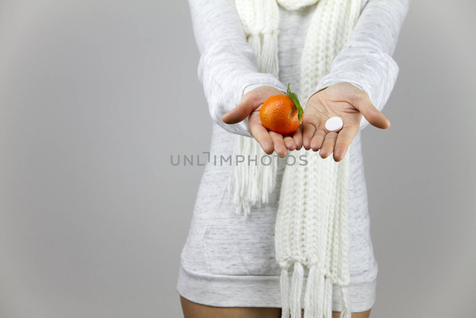 Vitamins from fruit or drugs? A sick young woman with a scarf on her neck shows a mandarin in her right hand and an aspirin on her left