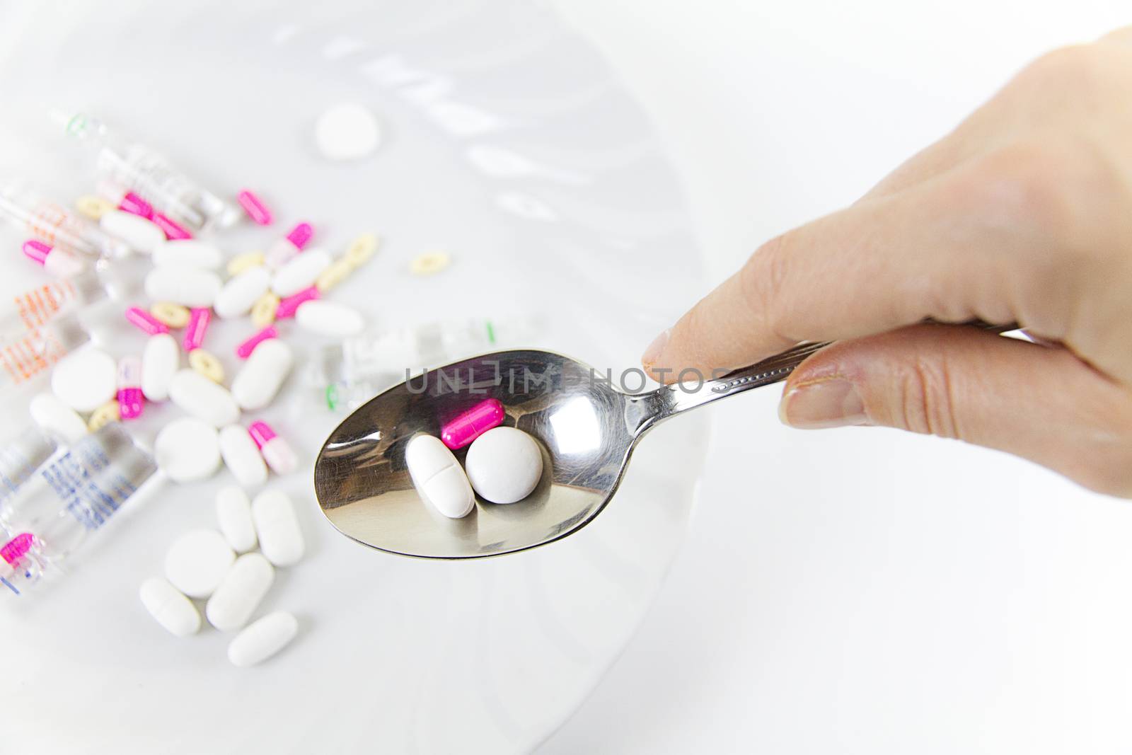 Addiction to medicines and drugs in modern times: a hand of a young woman with a spoonful of tablets taken from a plate full of medicines on white table and background