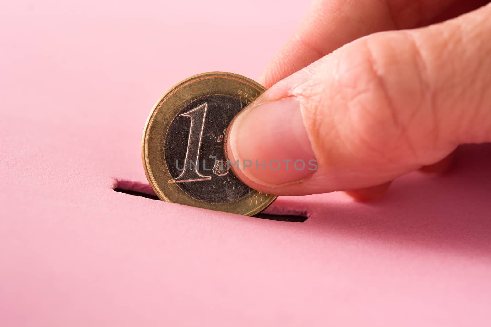 Hand putting one euro coin into a moneybox on pink background