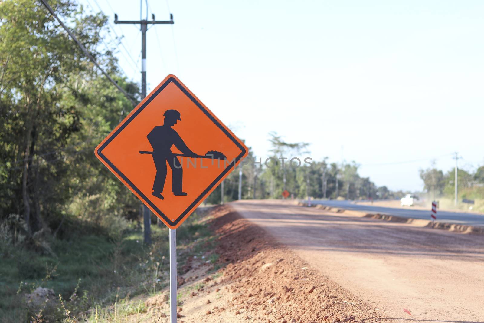 The orange sign shows a symbol with the image of a person holding a shovel on the sign installed on the side of the construction road. Warning sign indicating road work is ahead.