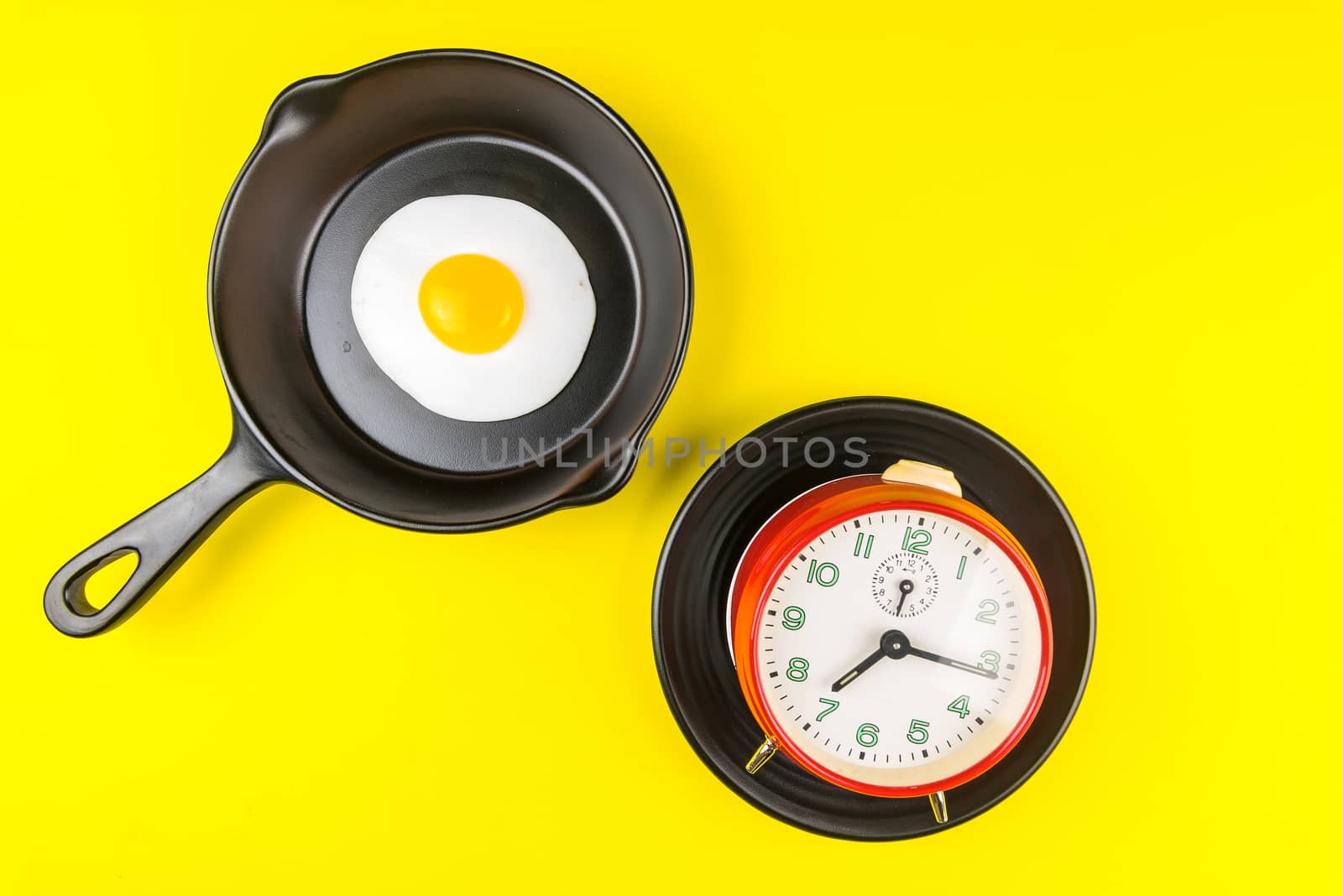 Frying pan with fried egg and alarm clock in a black plate isolated on a yellow background viewed from above.Concept of breakfast at the race against time.