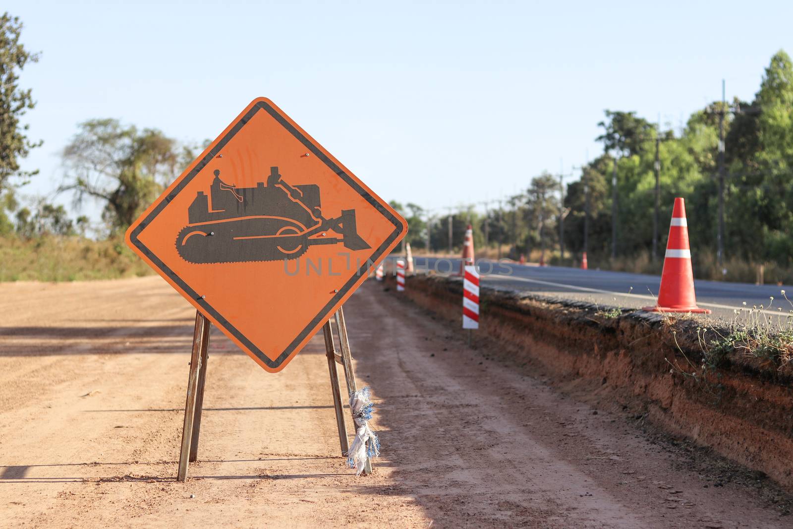 The orange sign shows the machine symbol being operating on the side of the road. Warning sign indicating road work is ahead. by Eungsuwat
