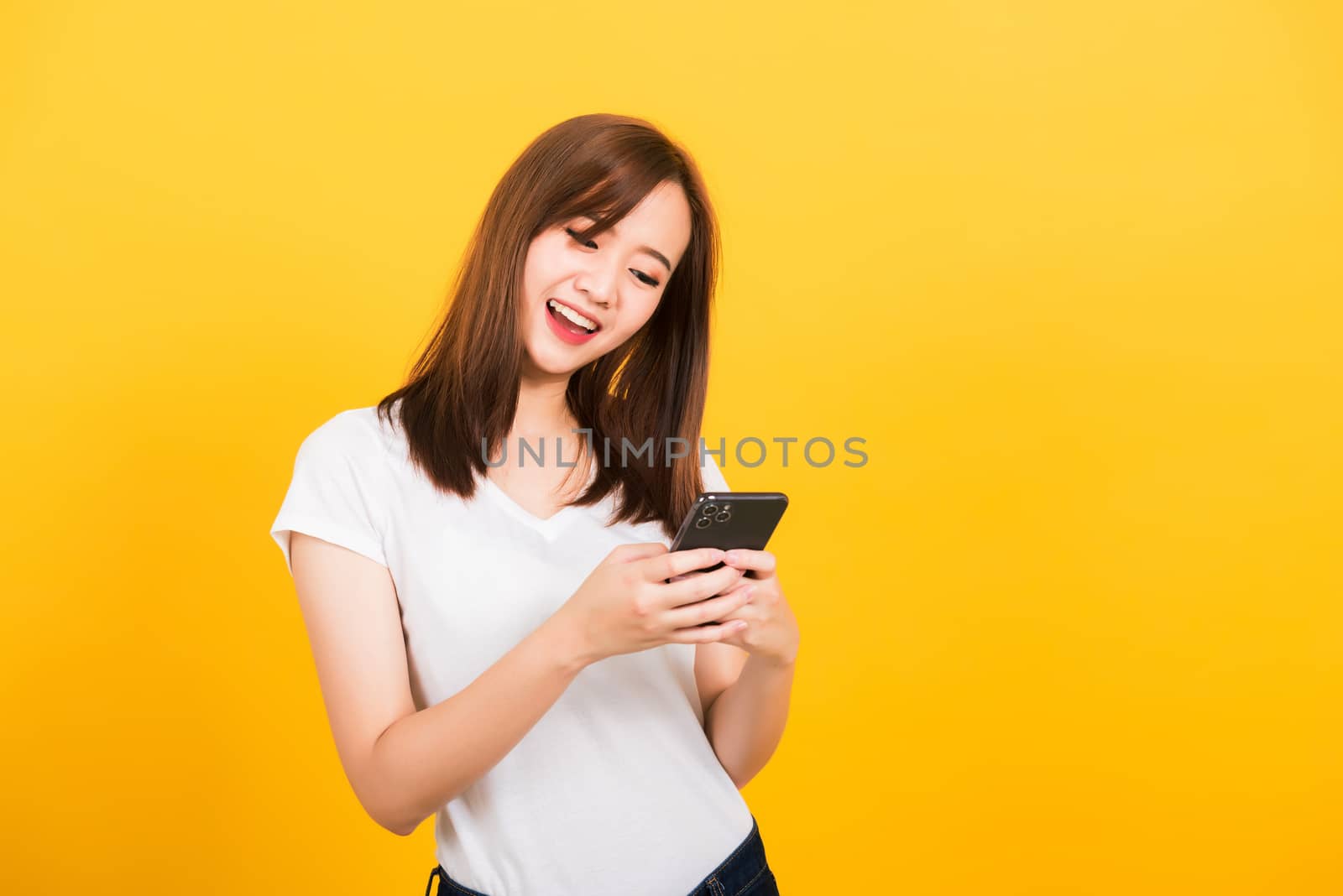 Asian happy portrait beautiful cute young woman teen smile standing playing game or writing message on smartphone looking to the phone isolated, studio shot on yellow background with copy space