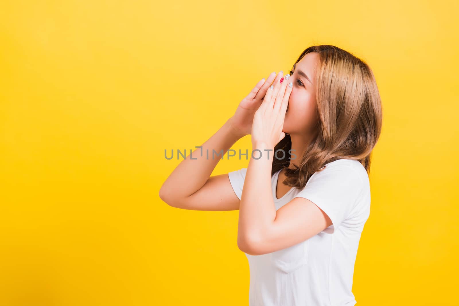 Asian Thai happy portrait beautiful cute young woman teen standing big shout out with hands next mouth giving excited positive looking to camera isolated, studio shot on yellow background with copy space