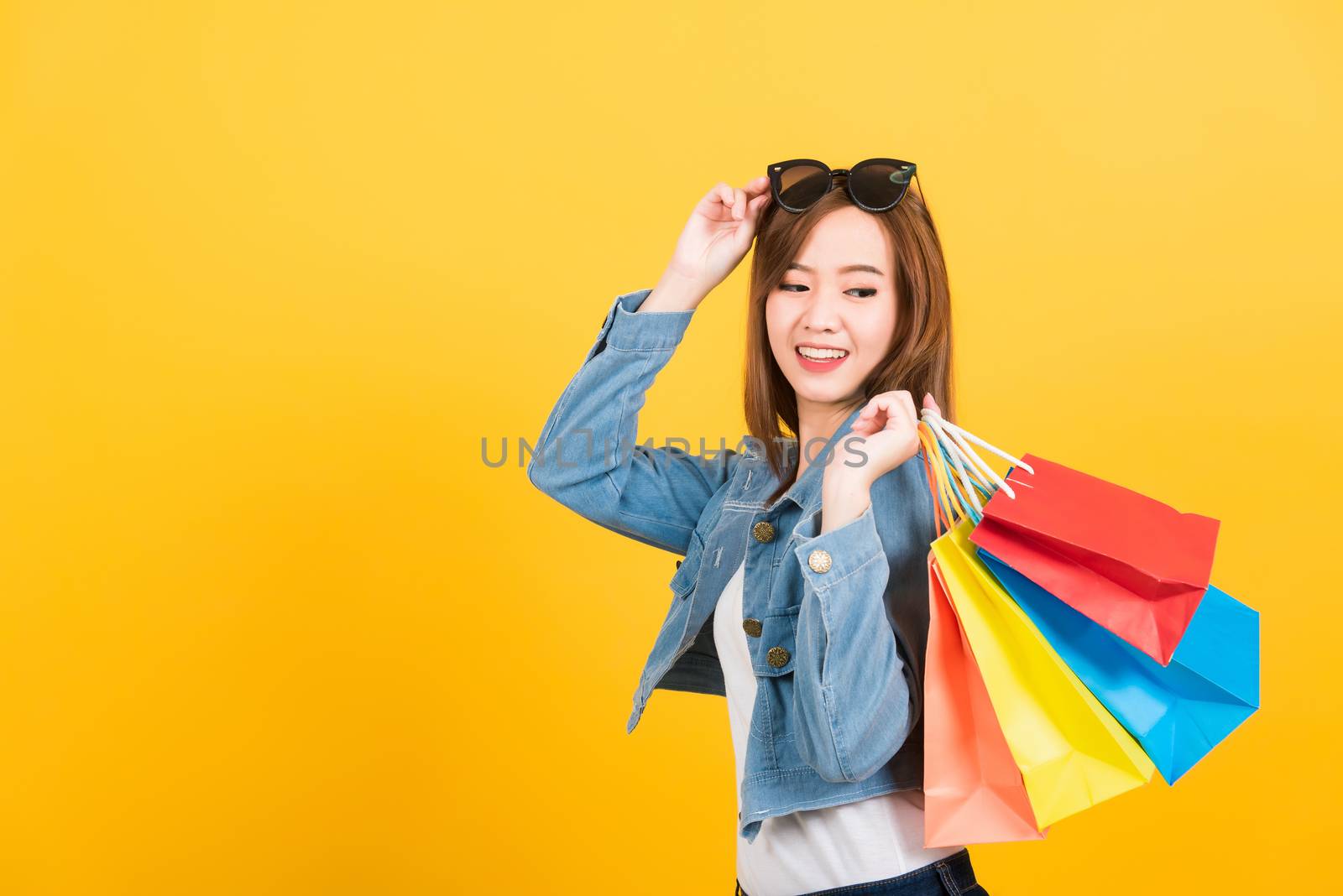 Asian happy portrait beautiful cute young woman teen smiling standing with sunglasses excited holding shopping bags multi color looking bags isolated, studio shot yellow background with copy space