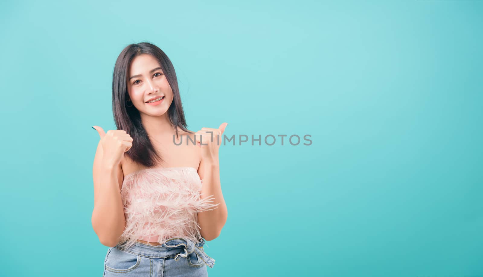 Asian happy portrait beautiful young woman standing smiling showing hand showing thumbs-up sign and looking to camera isolated on blue background with copy space for text