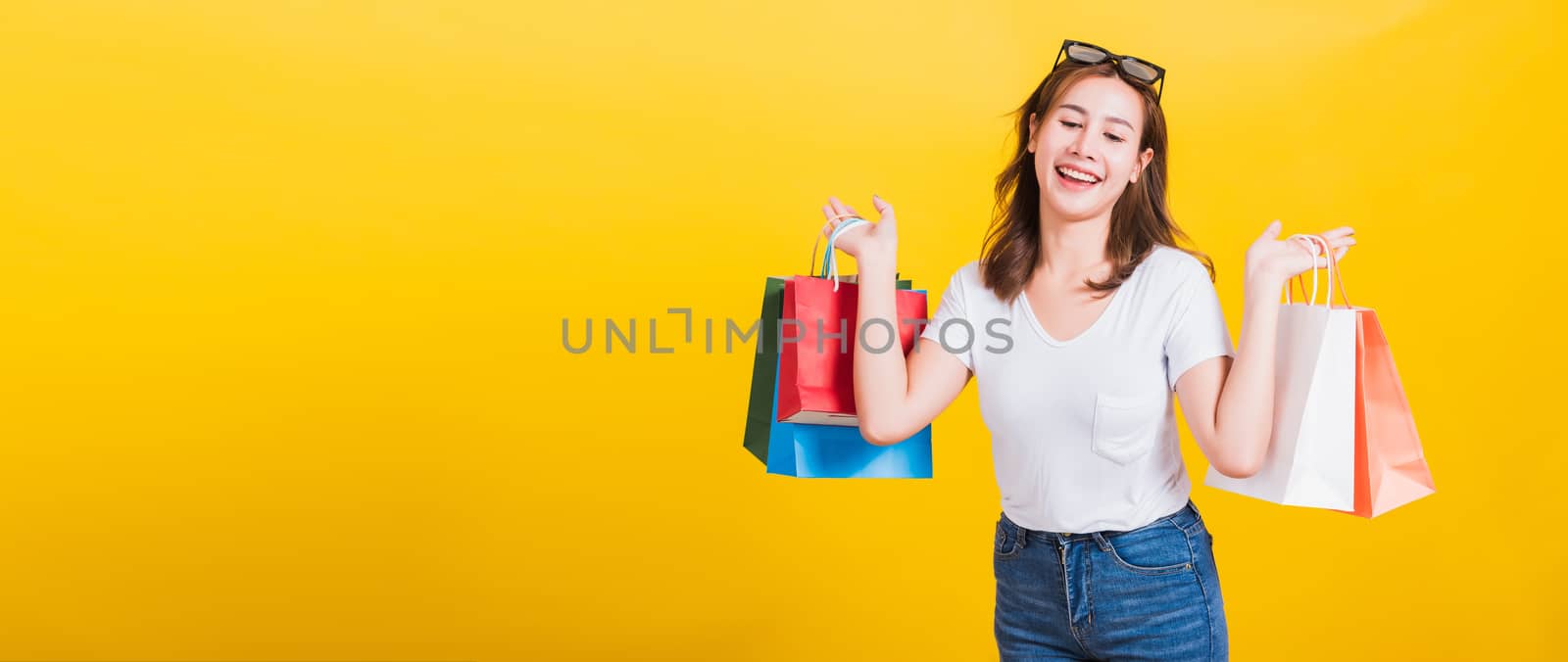 Asian Thai portrait happy beautiful cute young woman smiling stand with sunglasses excited holding shopping bags multi color looking down, studio shot isolated yellow background with copy space