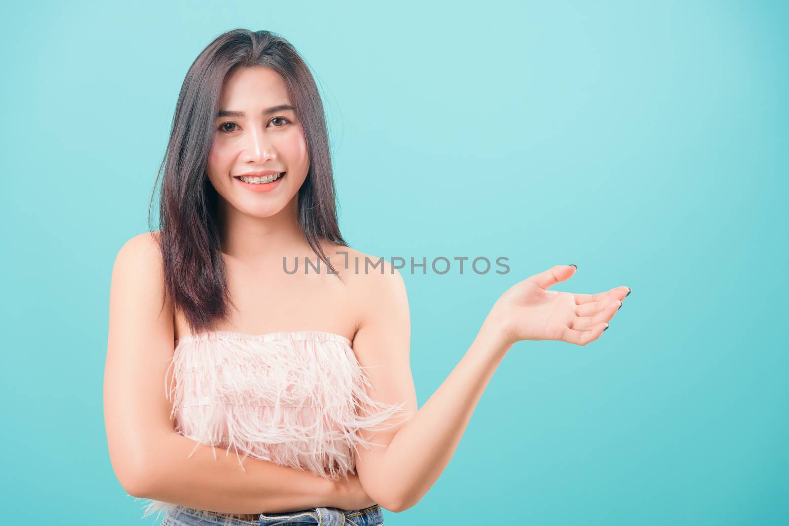 Asian happy portrait beautiful young woman standing her showing hand something near body and looking to camera isolated on blue background with copy space for text