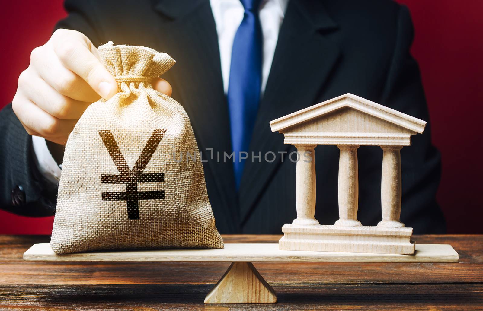 A man puts a Yen yuan money bag on scales opposite to building of government, bank, university. Budget and funding for normal functioning. Deposits and loans. Lobbying interests. Payment of taxes.