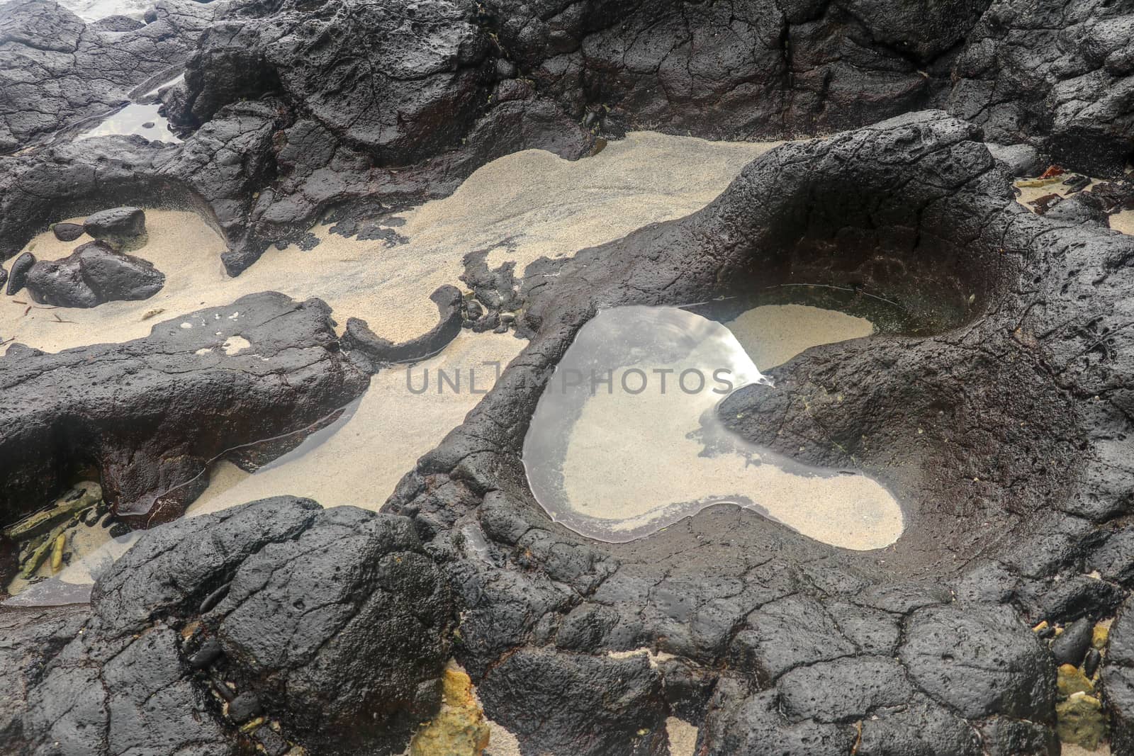 Depressions in a rock with white sand at the bottom, flooded with sea water. Small ponds excavated in lava bed on Indian Ocean coast, Bali, Indonesia. The tide dug out small lakes in the black rock.