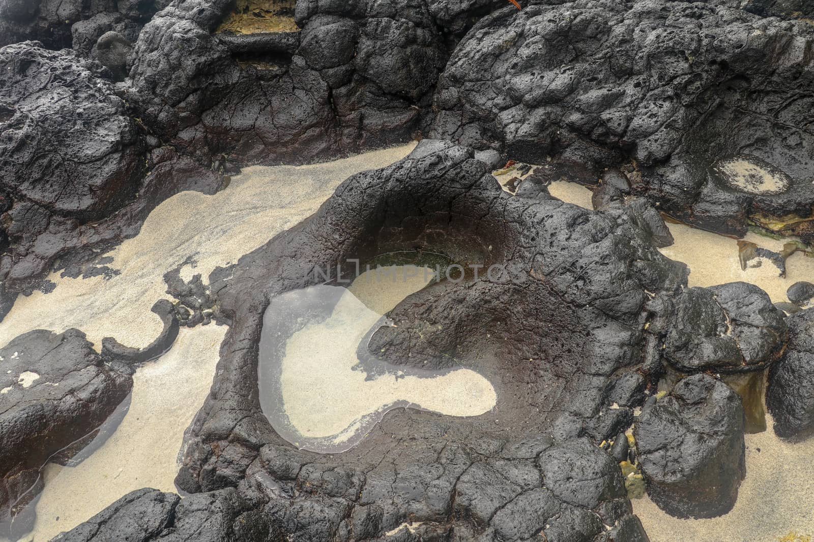 Depressions in a rock with white sand at the bottom, flooded with sea water. Small ponds excavated in lava bed on Indian Ocean coast, Bali, Indonesia. The tide dug out small lakes in the black rock.