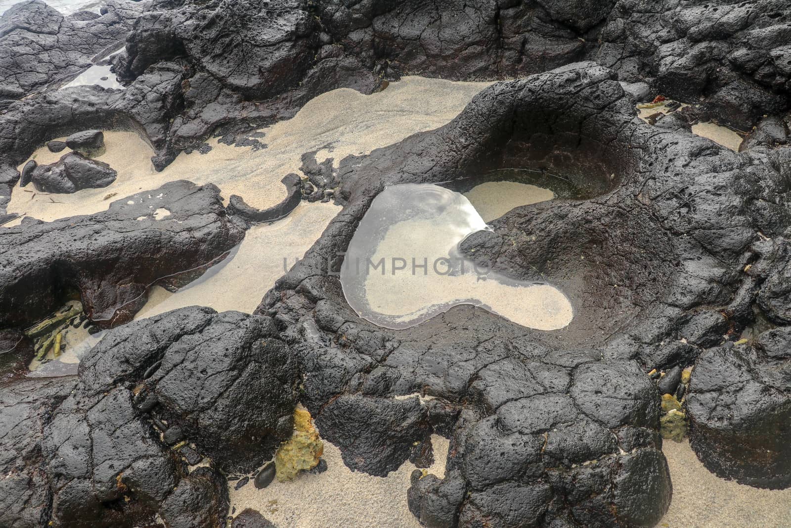 Depressions in a rock with white sand at the bottom, flooded with sea water. Small ponds excavated in lava bed on Indian Ocean coast, Bali, Indonesia. The tide dug out small lakes in the black rock.
