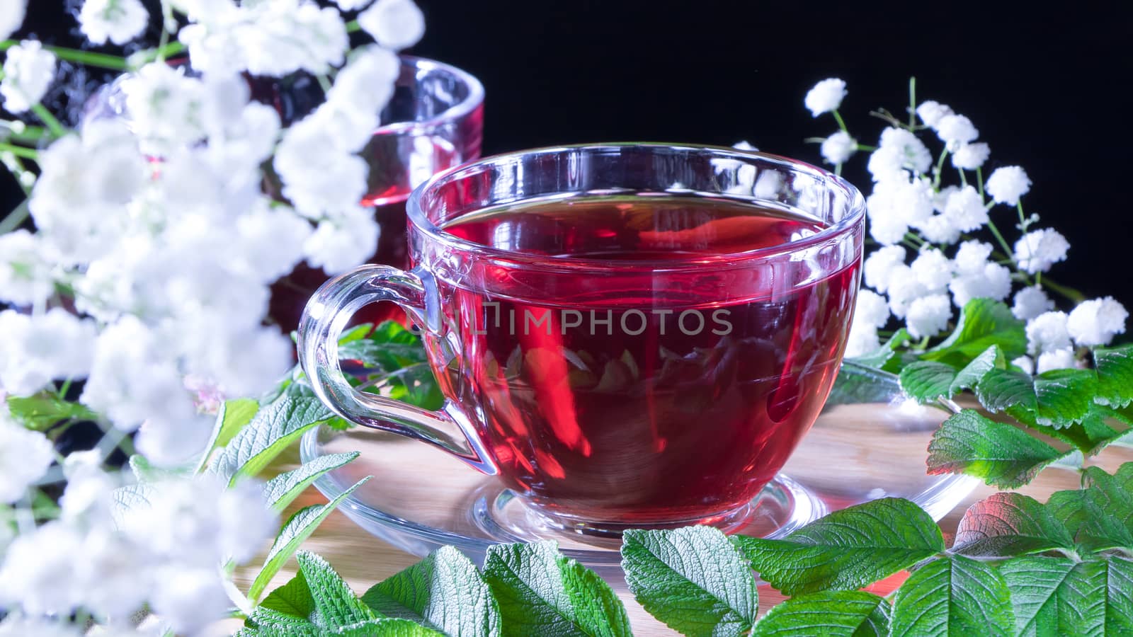 A mug of red tea in white hibiscus flowers and green leaves of medicinal tea on a wooden stand.Zen tea ceremony. Photo of red herbal Indian healing tea. Elegant mugs with a relaxing and tonic drink